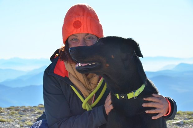 Une femme au bonnet orange enlace un grand chien noir devant les montagnes.